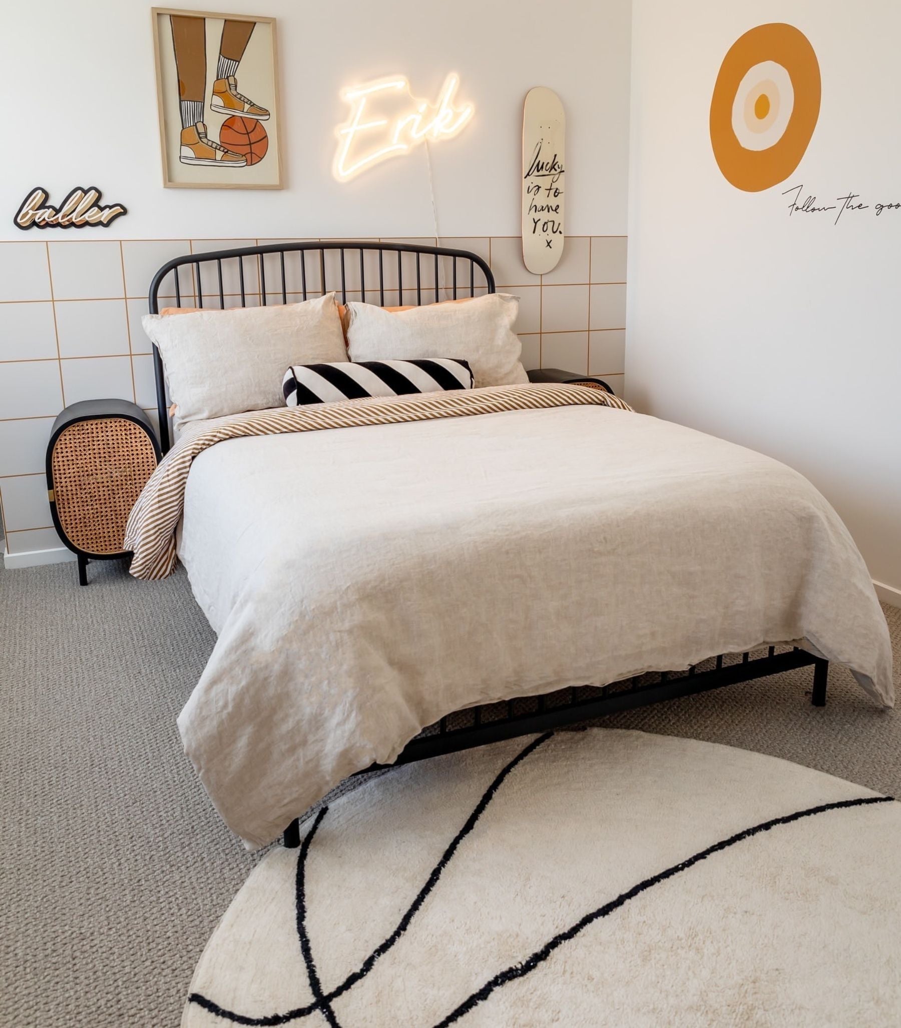Boys bedroom with black iron bed frame, linen bedding, rattan bedside table and circular basketball-style rug. Wall styled with neon name sign, skateboard and framed sneaker art in warm mustard and neutral tones.