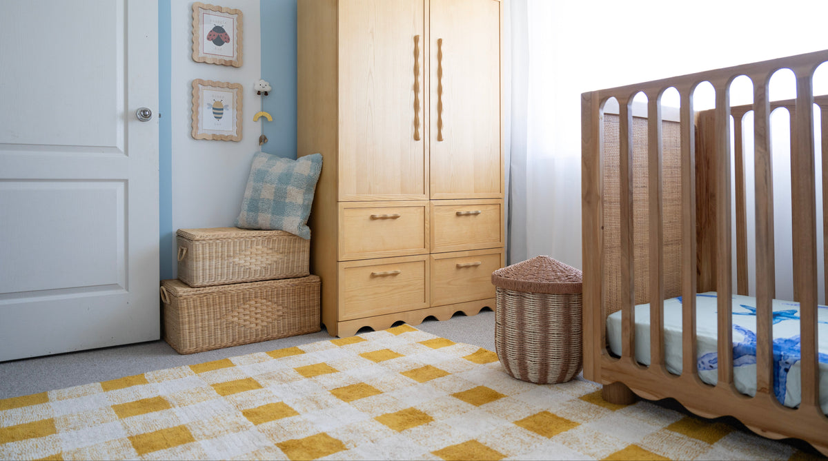 A styled nursery room featuring a light natural wood wardrobe, a wooden crib, a yellow and white checkered rug, and a blue and beige patterned pillow on a wicker bench.