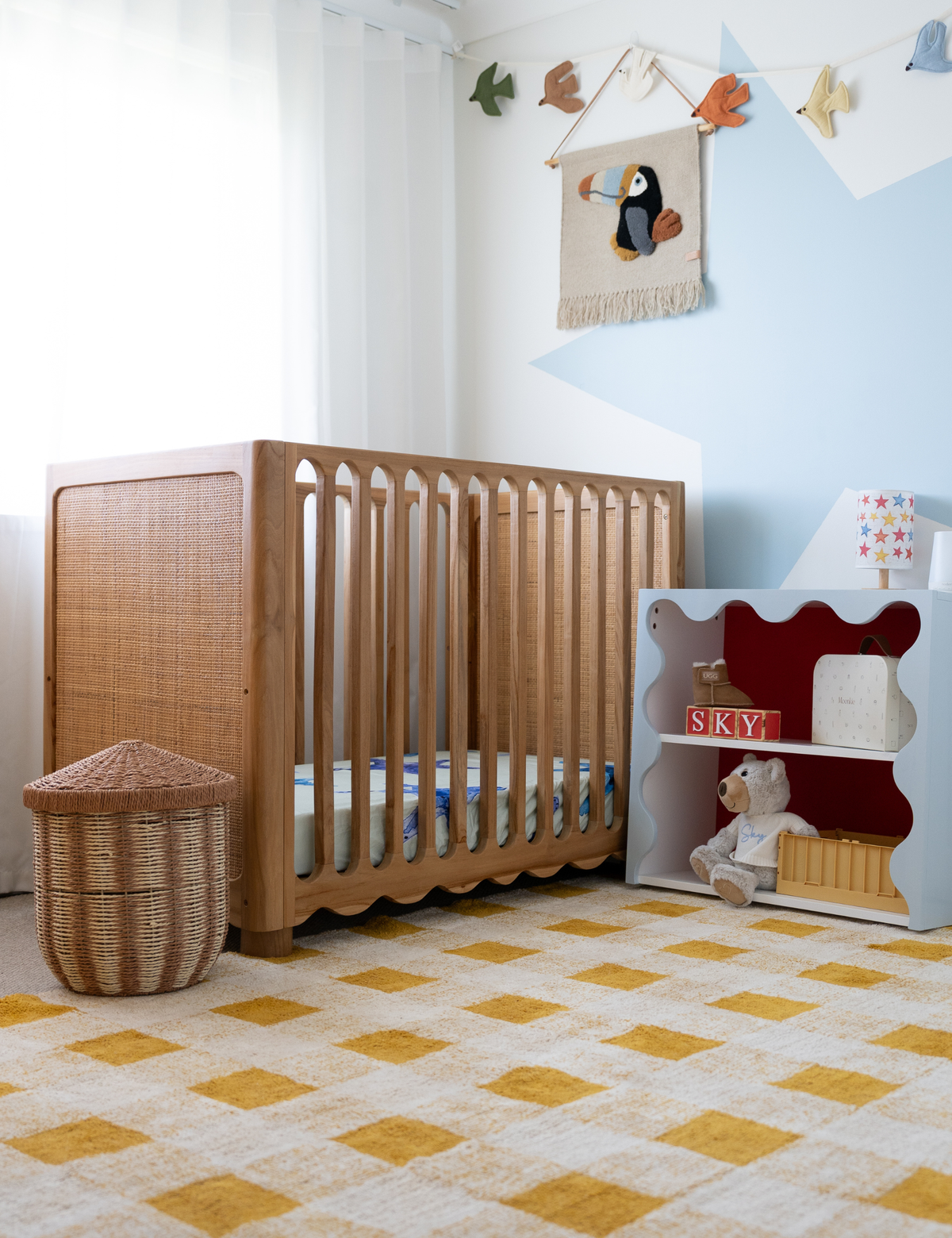 A wooden crib with a woven rattan headboard and scalloped bottom rail, styled in a bright nursery with a yellow-and-white checkered rug, a white storage shelf, and a penguin wall hanging.